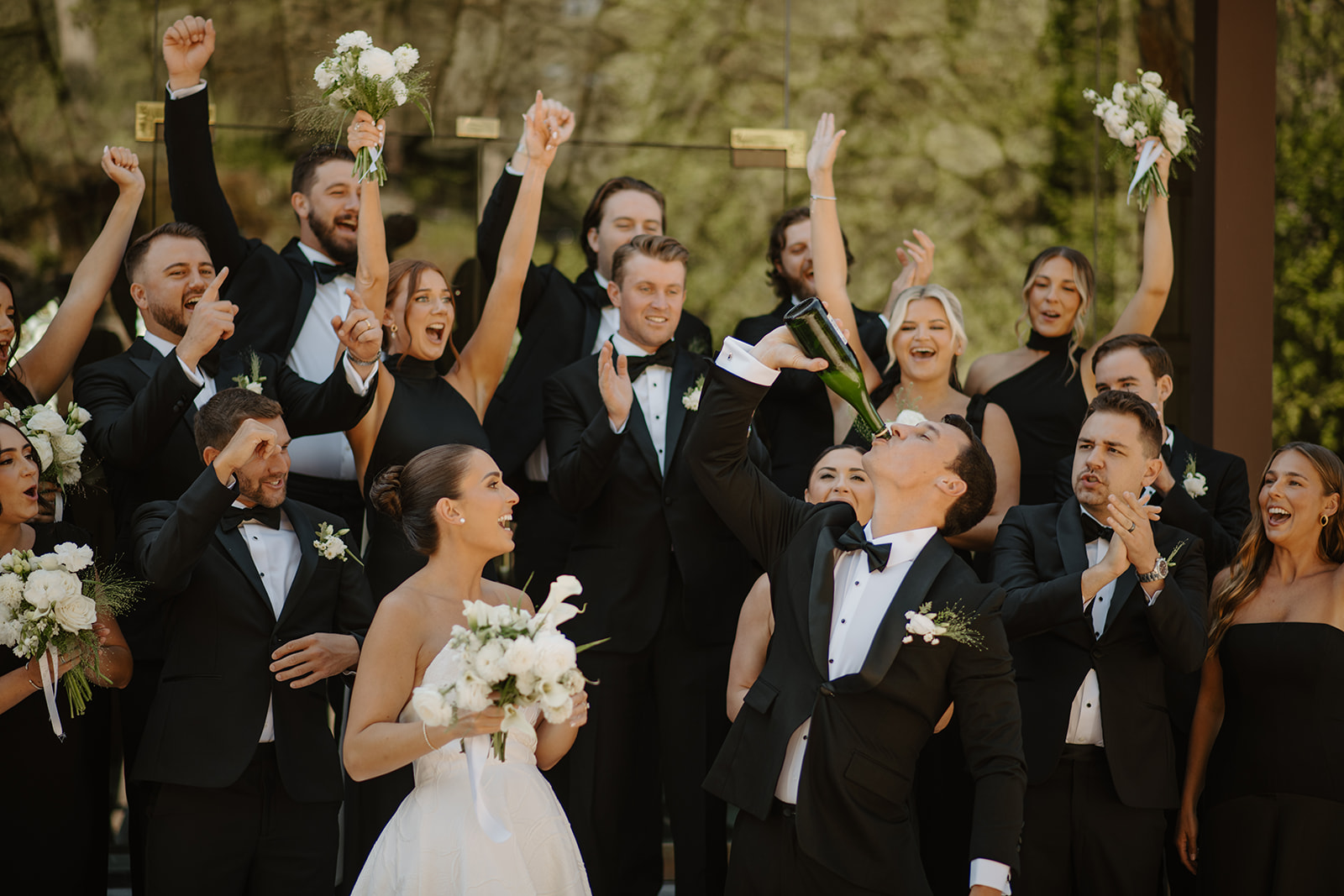Groom drinking straight from a champagne bottle while the bride and wedding party cheer him on.